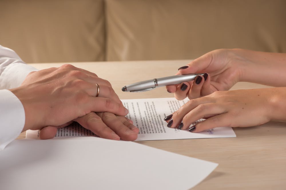 Two hands, one passing a pen over a document on a desk—suggestive of an agreement signing within an office setting.