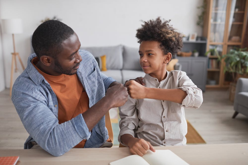 A man and a young boy are fist bumping as a gesture of agreement or solidarity, sitting at a table in a cozy living room.