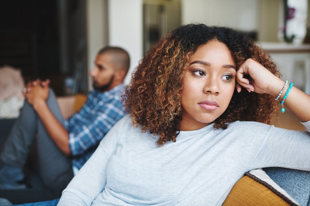 A woman appears contemplative, resting her head on her hand, while a man sits in the background out of focus, within a domestic setting.