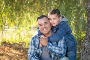 A man and a young boy in denim outfits embrace, smiling in a sunlit, leafy park.