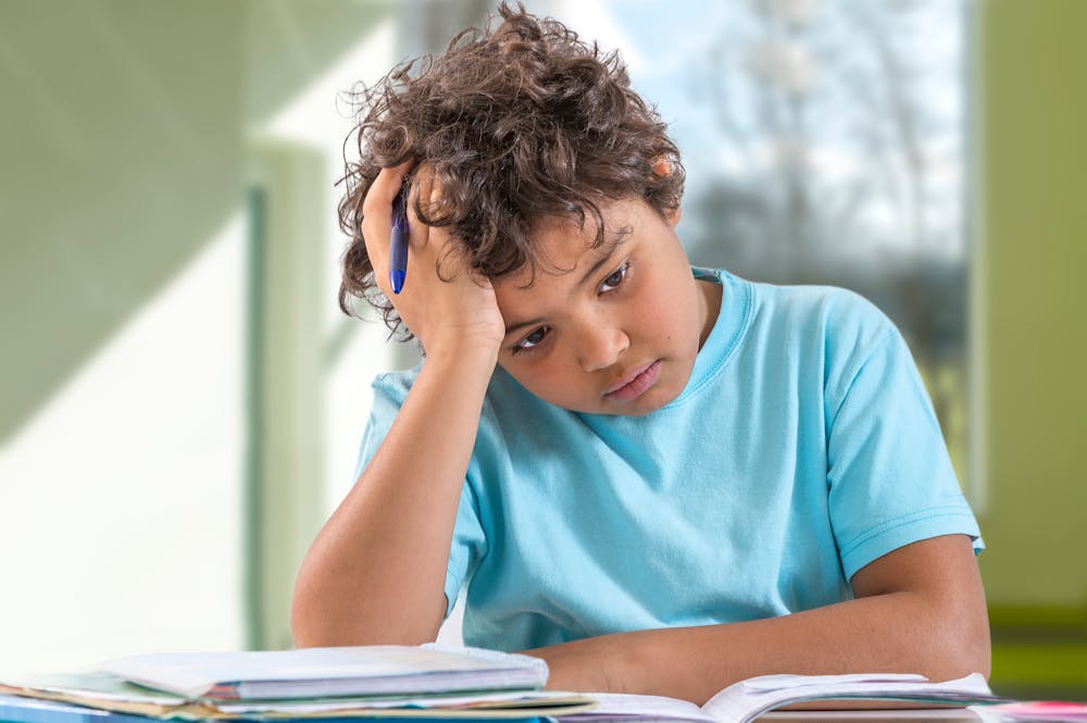 A child in a light blue shirt appears frustrated or thoughtful while studying books at a desk, against a blurred classroom background.