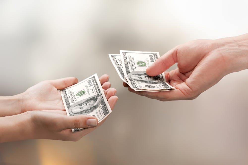 Two hands exchanging US $100 bills against a blurred background, depicting a financial transaction or payment.