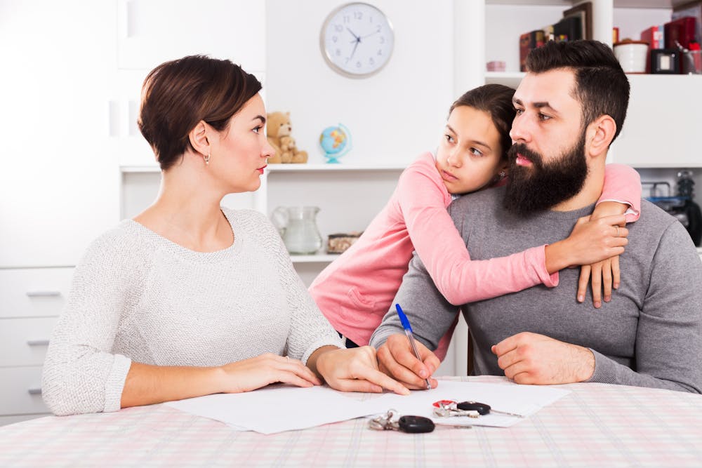 A family of three is engaged in discussion at a kitchen table. A man embraces a child while conversing with a woman, with papers and keys on the table.