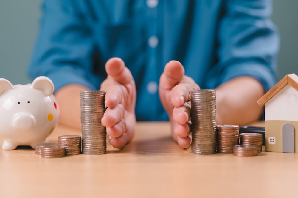 A person's hands are protecting stacked coins, with a piggy bank and a house model nearby, symbolizing savings and investment in real estate.