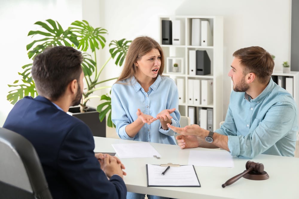 A woman gestures emphatically during a discussion with two men at a table with documents and a gavel, suggesting a dispute or negotiation in an office setting.