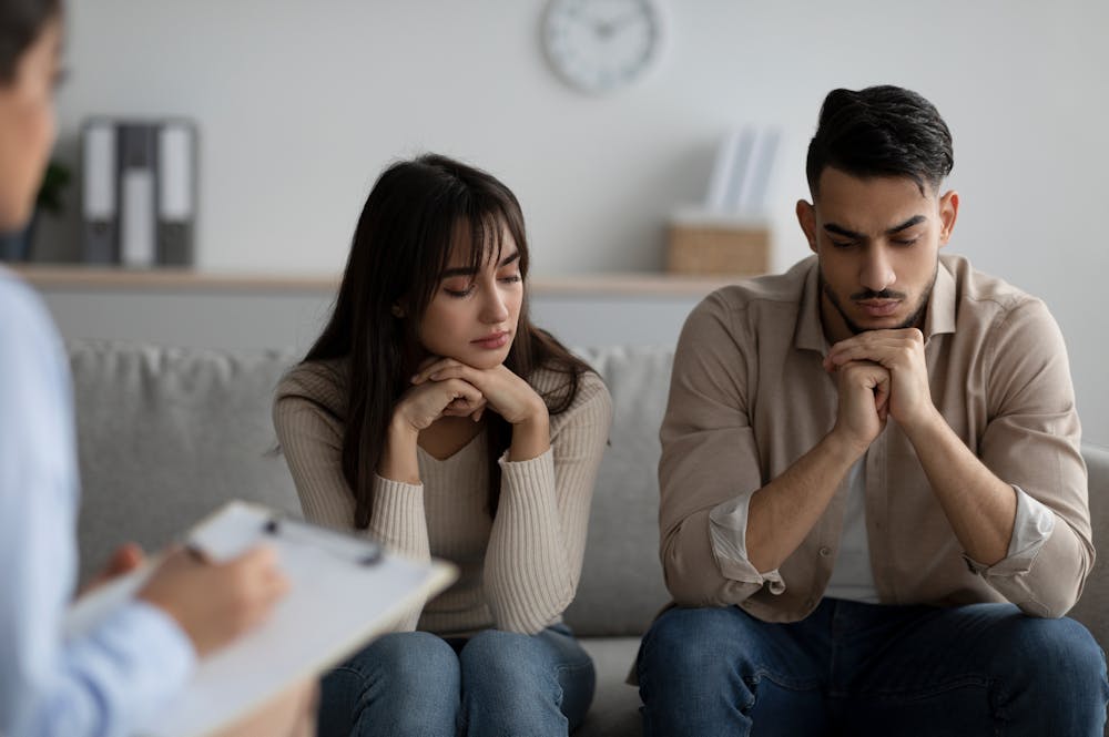 A man and woman sit side by side, appearing contemplative or worried, while another person takes notes on a clipboard, suggesting a counseling or consultation setting.