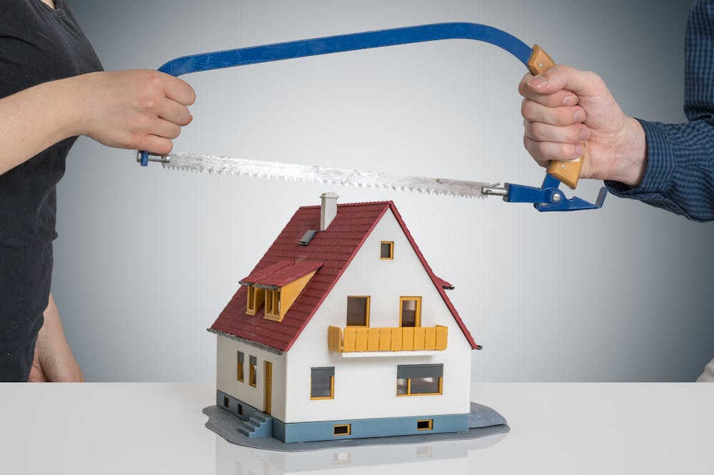 Two hands hold a saw over a small model house, implying cutting it in half, against a grey background.