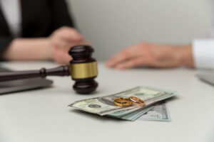 A gavel rests beside two gold wedding rings and a stack of cash on a table, implying a legal issue involving marriage finances, with a partial view of a person's hands in the background.