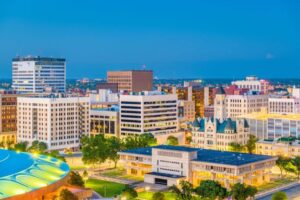 A cityscape at dusk with illuminated buildings against a twilight sky, featuring a mix of modern and historic architecture, and a clear foreground park area.