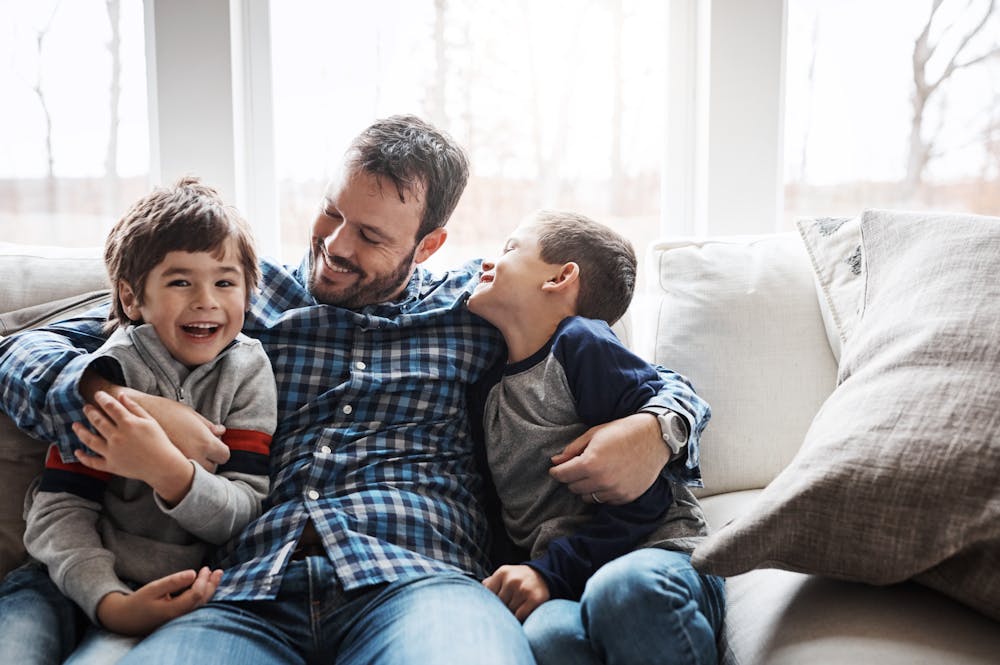 A man and two boys are laughing and cuddling on a sofa, in a well-lit room with large windows.