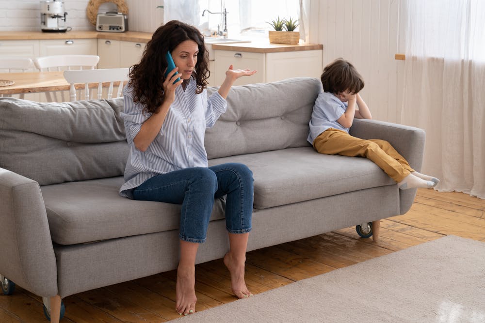 A woman talks on a phone, gesturing perplexedly. A child sits at the other end of the sofa, head buried in hands. They're in a cozy, well-lit living room.