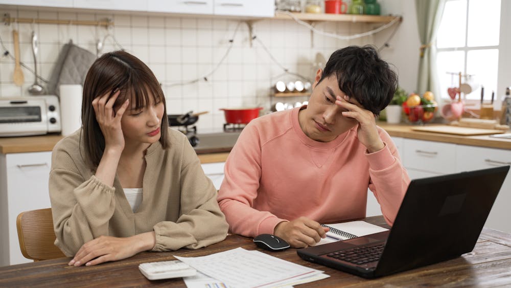 Two individuals appear stressed while looking at a laptop in a kitchen setting, surrounded by household items and paperwork on the table.