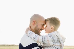 A man and a young boy are smiling and touching foreheads affectionately in a bright, open field. They appear joyful and are embracing closely.