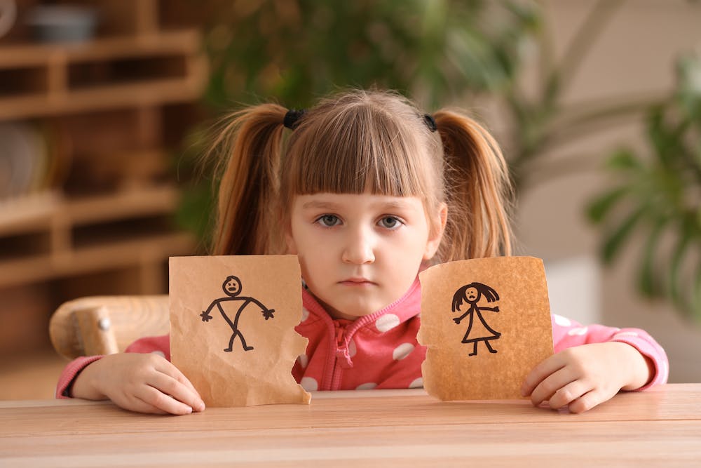 A young girl with pigtails holding two drawings of stick figures, looking serious, sitting at a wooden table, with a blurred indoor background.