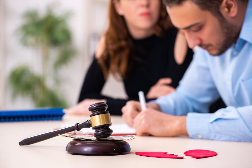 A man and woman, sitting at a table with a judicial gavel and broken heart, evoke a divorce context. The man is signing documents, possibly related to the proceedings.