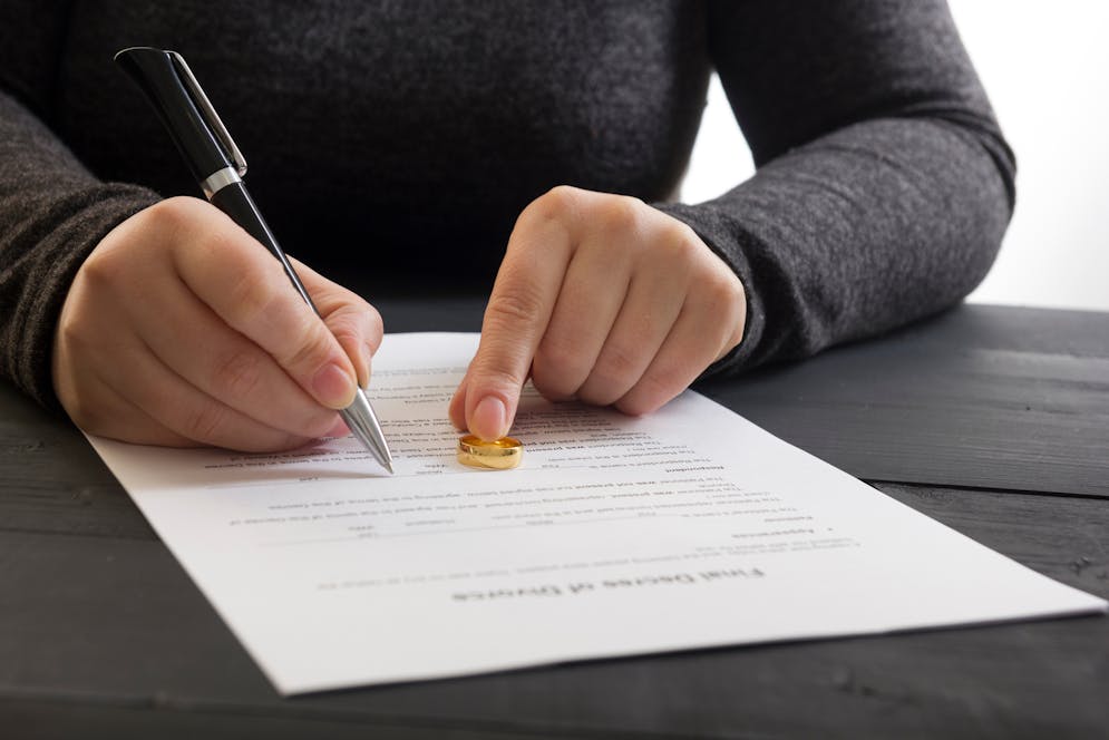 A person places a wedding ring on a document, signaling a divorce action, with a pen in hand. The document rests on a dark wooden table.