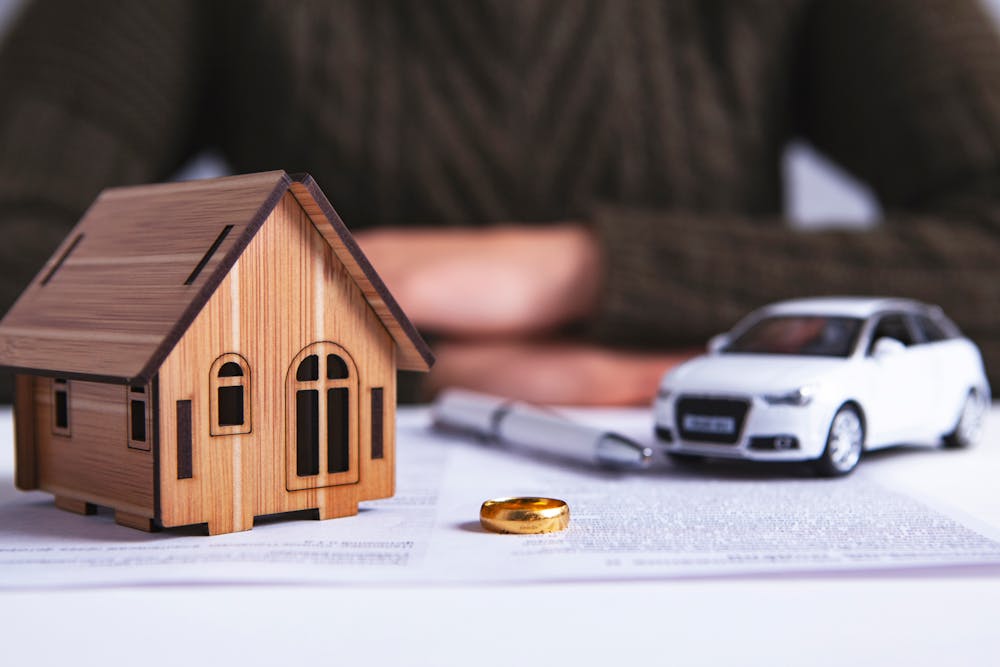 A wooden model house and a white car model sit near folded hands, with a gold ring and documents on a desk, suggesting themes of ownership and investment.