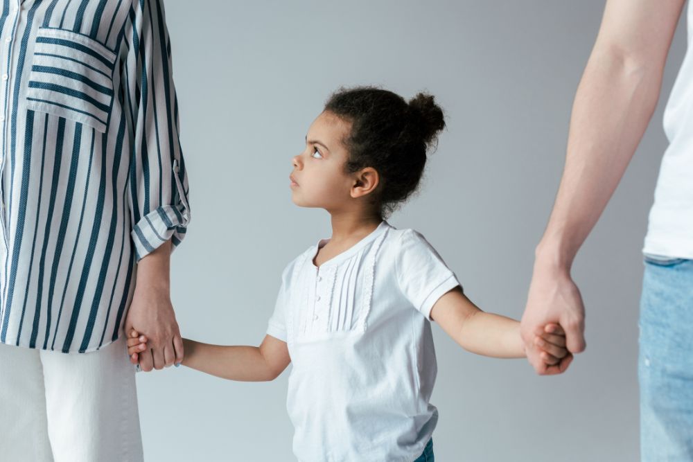 A young girl holding hands with two adults, looking up at one, against a plain background.