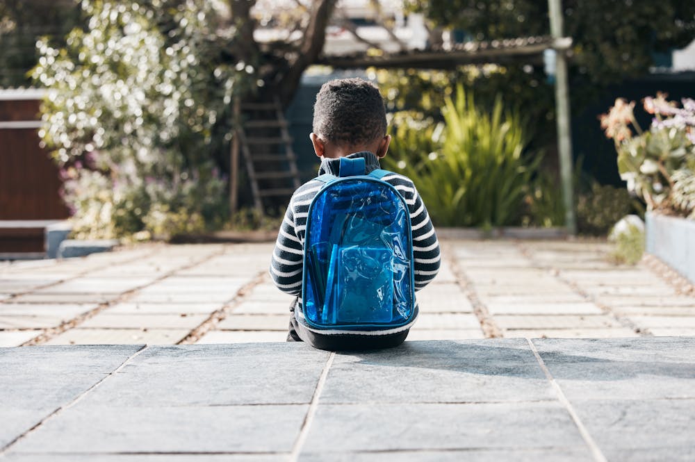 A child with a blue backpack sits alone on a stone pathway, surrounded by garden foliage.