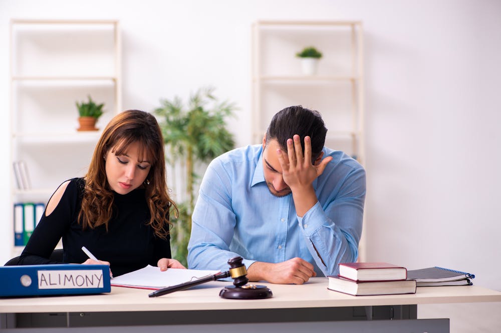 Two people appear stressed, sitting at a desk with a folder labeled "ALIMONY" and a gavel, suggesting a legal or divorce-related context.