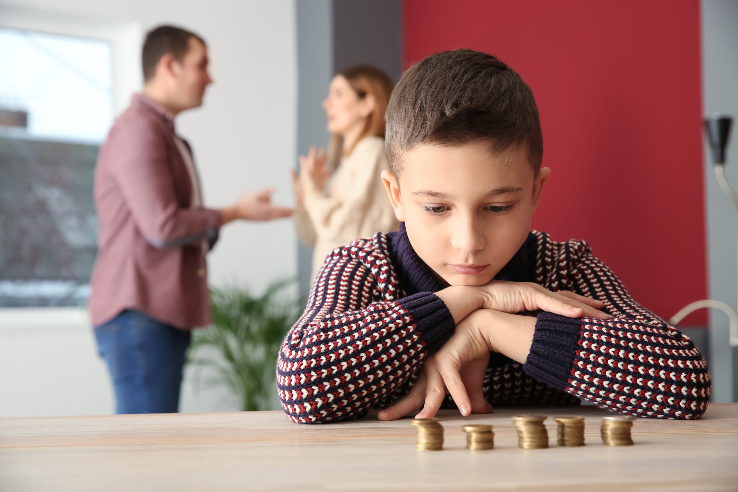 A young boy looks at coins on a table, appearing thoughtful, with a blurred couple arguing in the background, suggesting family financial difficulties.