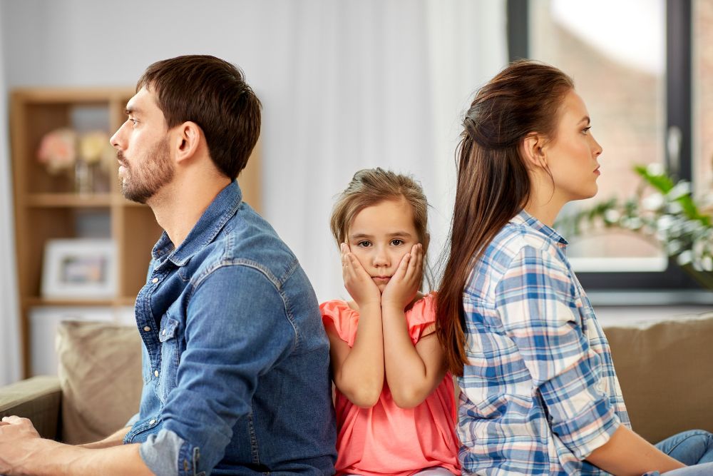 A man, woman, and child sit on a couch, facing opposite directions, looking upset, in a brightly lit living room.