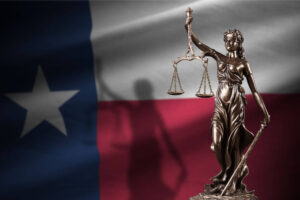 A bronze statue of Lady Justice holding scales set against the Texas state flag background.