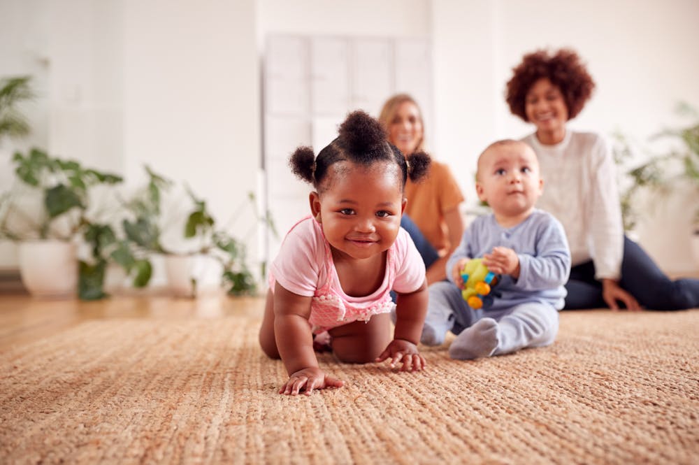 A toddler crawls on a carpet towards the camera, with two women and another baby seated behind in a bright, plant-adorned room.