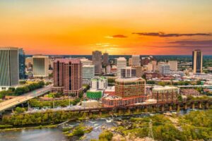 A skyline displaying numerous buildings at sunset, with a river flowing in the foreground and verdant trees peppered throughout the urban landscape.