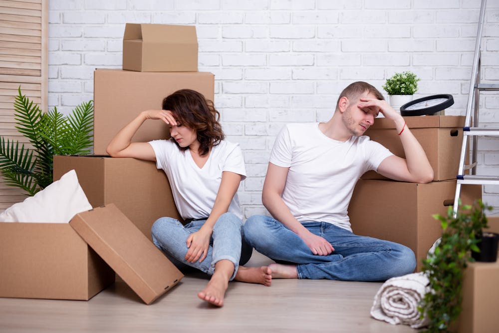 Two people sit exhausted among moving boxes in a white-walled room, suggesting the action of relocating or unpacking.