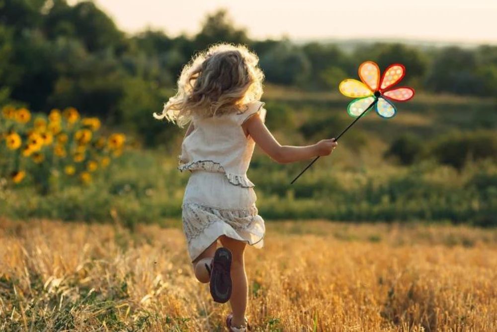 A young child runs through a sunlit field, holding a colorful pinwheel, with sunflowers and trees in the background.