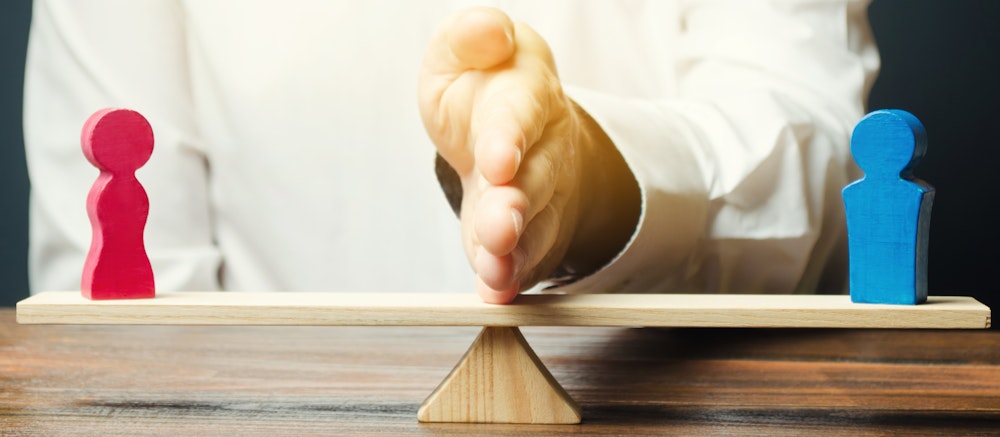 A person's hand tipping a seesaw, balancing red and blue figurines, suggesting a concept of equality or decision-making in an office-like setting.