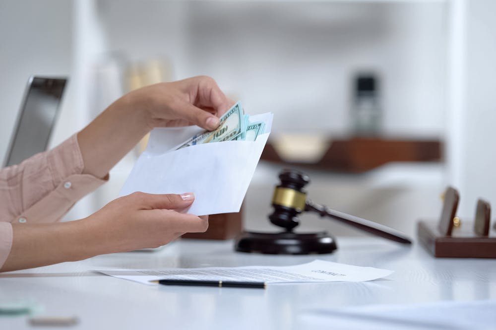 Hands exchanging money from an envelope, implying a transaction; focused on the foreground with blurred gavel and legal books in the background, suggesting a legal or judicial setting.