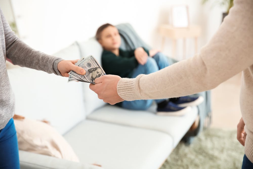 Two individuals exchange money, with one handing cash to another, against a blurred background of a person on a couch, suggesting a casual indoor setting.