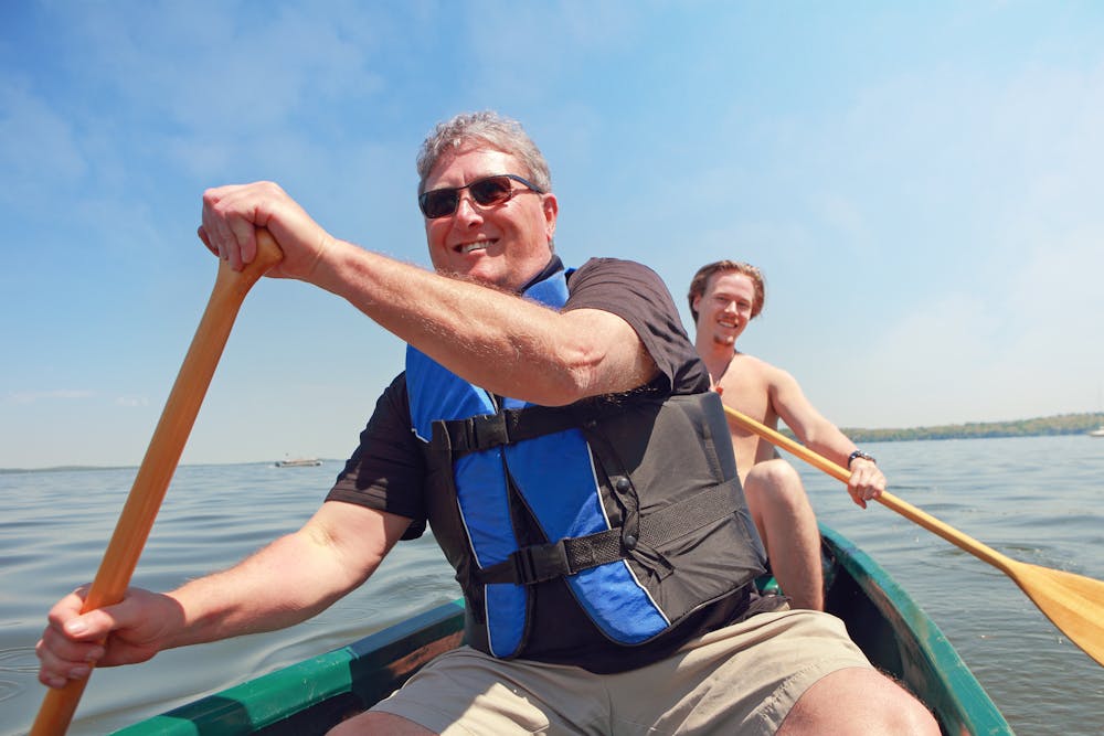 Two men are paddling in a canoe on a calm lake with clear skies. The older man in the foreground is wearing sunglasses and a life jacket.