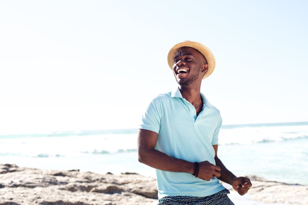 A joyful man in a straw hat and casual summer clothes is laughing by a sunny beach with waves in the background.