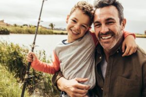 A smiling man and a boy, possibly father and son, hugging and holding a fishing rod by a lake, suggesting a bonding activity in a natural setting.