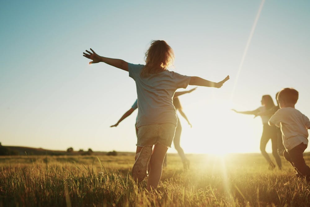 Children play joyously in an open grassy field during sunset, running with outstretched arms, backlit by the warm glow of the sun. No text is present.