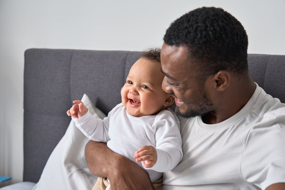 A smiling baby sitting with a man who is looking at the baby with affection, seated on a gray couch in a light-colored room.