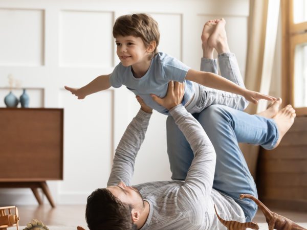 Joyful young man father lying on carpet floor, lifting excited happy little child son at home. Full length carefree two generations family having fun, practicing acroyoga in pair in living room.