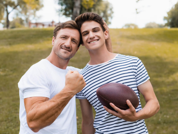proud father showing clenched fist and standing near teenager son with rugby ball