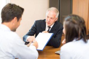 Three individuals in a business meeting, one presenting a document to sign, in an office setting.