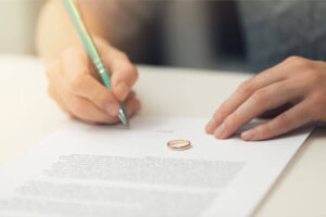 A person signs a document near a gold wedding band, likely indicating marriage dissolution in a calm, indoor setting. No visible text.