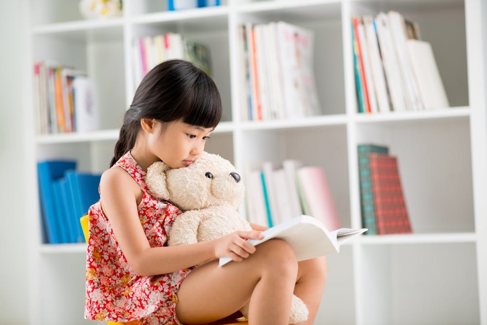 A young girl is reading a book, hugging a teddy bear, with bookshelves in the background.