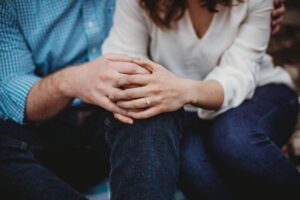 Two people, likely a couple, are sitting close together with their hands clasped on their laps, showcasing a diamond ring on the woman's finger, suggesting engagement.