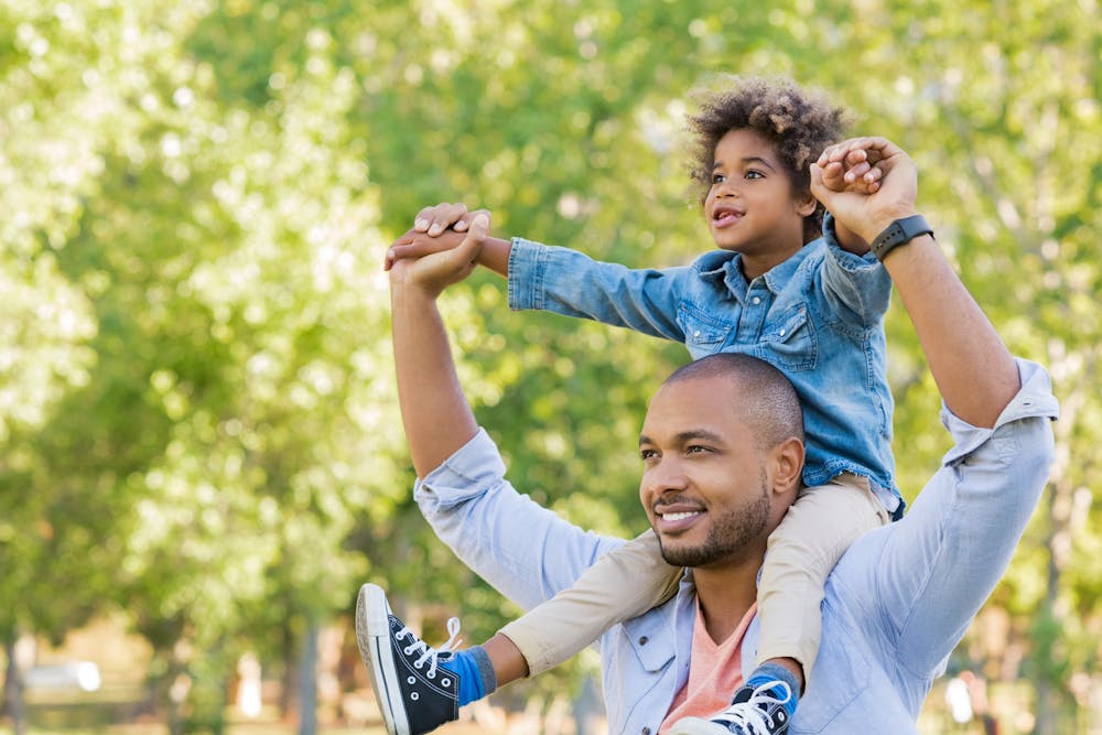 A smiling man carries a child on his shoulders in a sunlit, tree-filled park. The child looks up and away, both raising their arms joyfully.