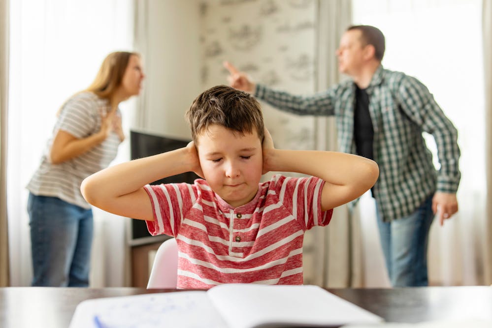 A young boy covers his ears with his hands, looking distressed, while behind him, two adults appear to be arguing in a domestic setting.