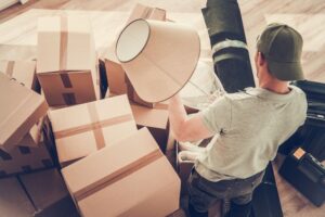 A person is packing items and furniture amidst numerous cardboard boxes in a room, suggesting moving out of a home.