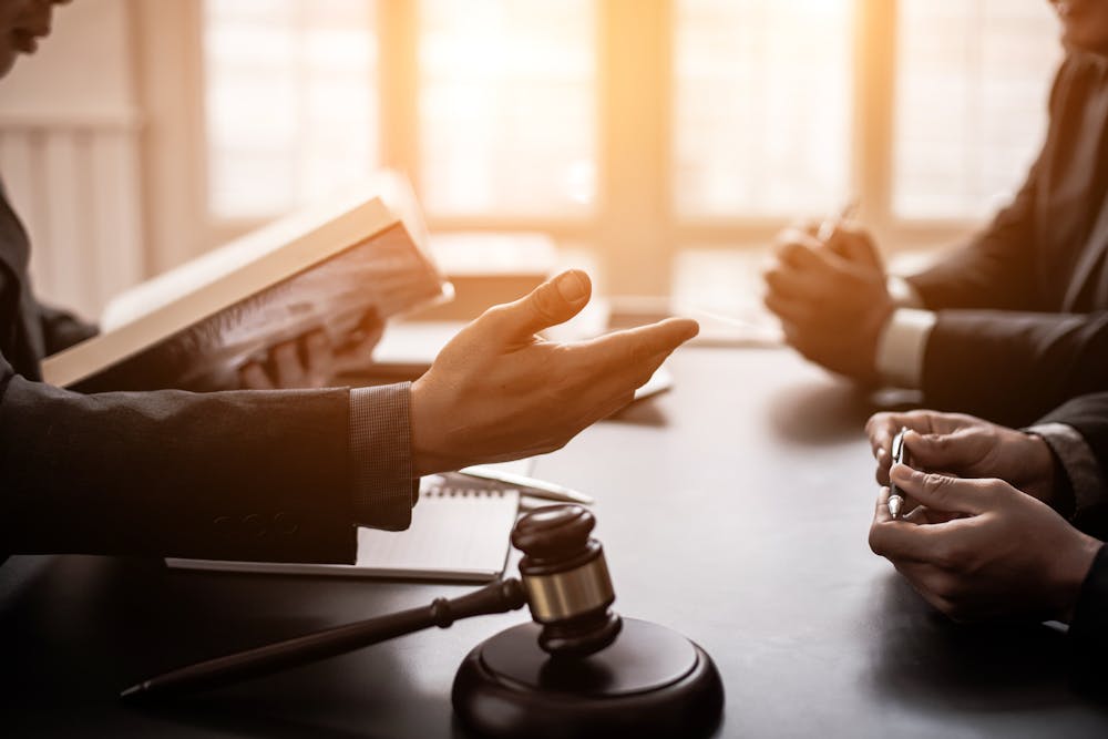 Two individuals are engaged in a conversation across a table, with one person gesturing with their hand. A gavel and a legal book are also visible, suggesting a legal context.