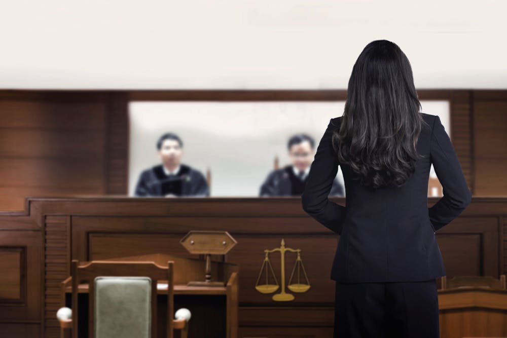 A person in a business suit stands facing a judge's bench in a courtroom, with the blurred image of a judge in the background.